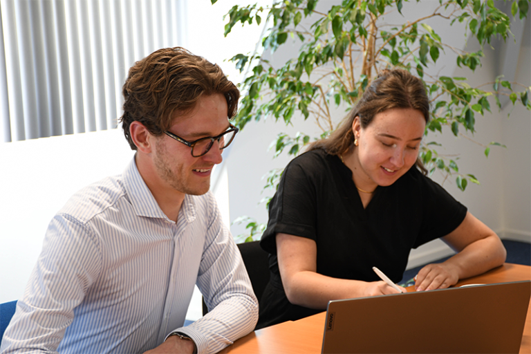 Foto van twee collega's naast elkaar aan tafel. De linker collega zit achter de laptop en de rechter schrijft op een notitieblok.